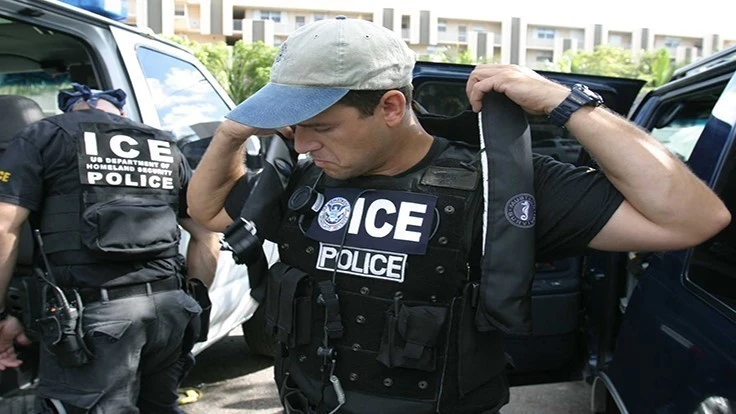 A man wearing a tan and blue baseball hat, black shirt and black police vest that says ICE Police.