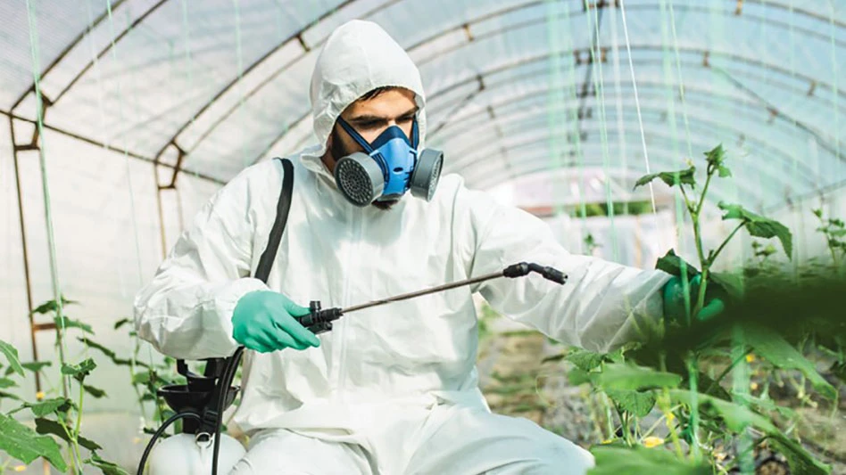 A man wearing a white suit, teal gloves and blue respirator holds a black spraying wand toward green plants as he squats in a greenhouse.