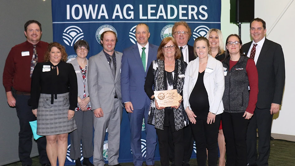 Eleven people pose for a photo in front of a dark blue backdrop that reads Iowa Ag Leaders in white capital letters. One person in the middle holds a wooden plaque in the shape of the state of Iowa.