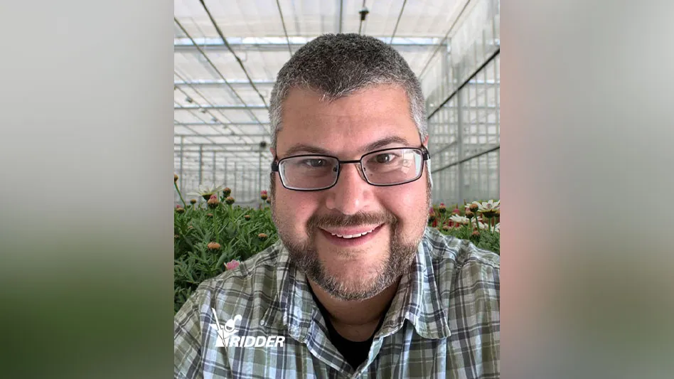 A smiling man with dark gray hair and a short dark gray beard wearing glasses and a green, brown and white plaid shirt takes a selfie in a greenhouse, with green plants visible behind him. In the bottom left of the photo is a logo that reads Ridder in white capital letters, with a graphic of a knight wearing a helmet and holding a lance to the left of the text.