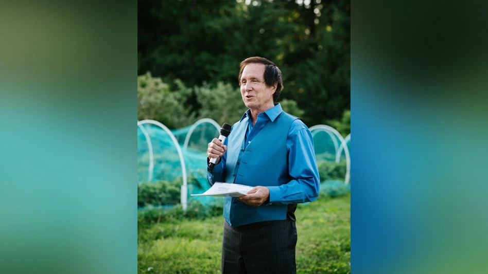 A smiling man with black hair wearing a blue shirt and vest holds a microphone in one hand and a piece of paper in the other. Green grass and trees are visible behind him.