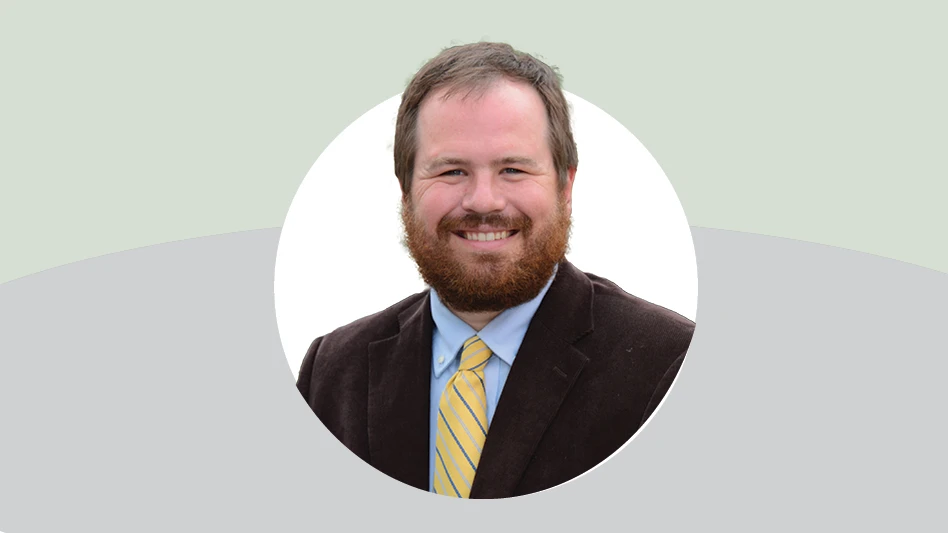 A smiling man with short brown hair and a short reddish brown beard, wearing a brown suit jacket, light blue dress shirt and yellow and blue striped tie. The background is gray, green and white.